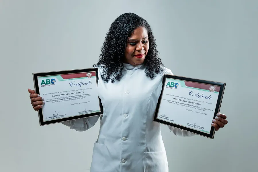 Aluna de jaleco branco com dois certificados emoldurados do Curso ASB