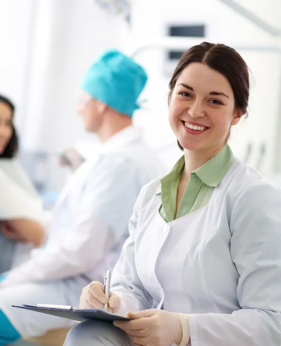 Profissional de saúde bucal sorrindo, de jaleco branco, com prancheta nas mãos numa clínica